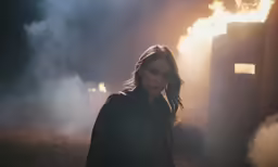 the girl looks back at the camera as she stands in front of a fire