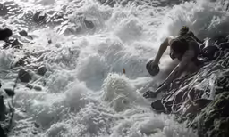 a person with a backpack reaches over rocks into the water