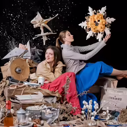 two women sit on a couch with the background scattered with items
