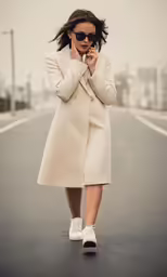 a woman in sunglasses, a coat and sneakers walking down a street