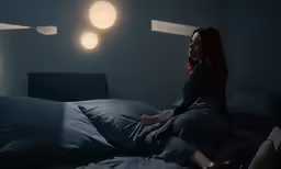 a woman in grey dress sitting on the edge of bed