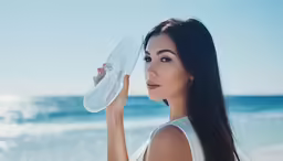 an elegant lady standing on a beach, posing with her hat and water bottle