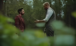 two men talking outside in a park surrounded by trees