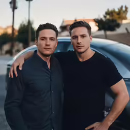 two young men pose together in front of a black car