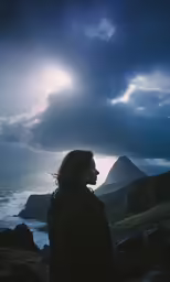 a woman with long hair standing next to the ocean under cloudy skies