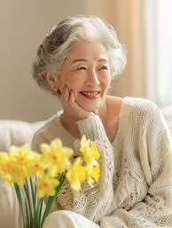 smiling old asian woman sitting on a couch and holding a flower,an older woman smiling at the camera with yellow flowers