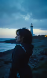 a woman looking away from the ocean with a lighthouse behind her