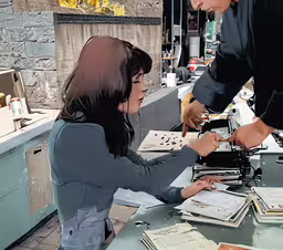 two people are looking at a table with papers,an older woman working on a piece of paper