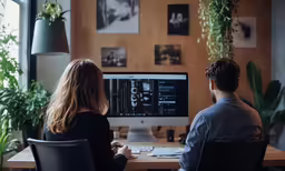 two people are sitting at a desk looking at a computer screen