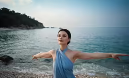 a woman in blue dress standing on the shore with her arms spread out