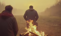 two men standing next to a campfire with the sun shining behind them