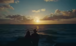 two people sit on rocks next to the ocean at sunset