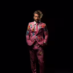 a bearded man in pink suit and tie,a man in a flowered suit stands in front of a black background