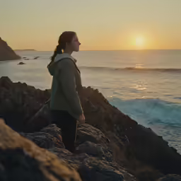 woman with back pack walking along rocky shoreline at sunset