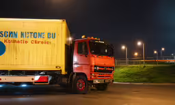 an orange truck driving down a street next to an empty field,an orange and red truck drives by light poles