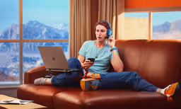 woman sitting on a sofa with headphones on her ears while listening to music and talking on her computer,a young woman sitting on a brown leather couch listening to music using her laptop