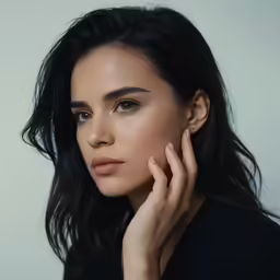 a woman with black hair and light brown eyes poses in a black dress with a neutral background