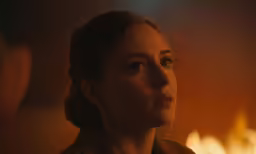 a woman stares into the distance with bright fire in the background