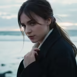 a woman with long hair and a jacket standing on a beach near water
