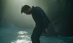 a young man in a dark shirt and a blue sweater stands on a rock over a river
