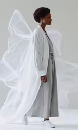 a woman in grey and white, with long organine veils, stands against a white backdrop