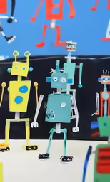 two robots sitting and standing in front of blue, yellow, red, and green paintings