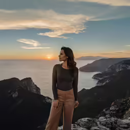 woman in wide legged pant and top stands on a cliff top overlooking the ocean