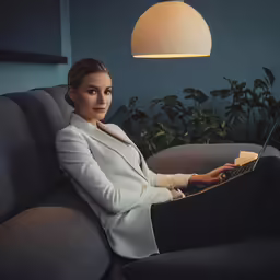 a woman in white jacket sitting on grey couch and looking at laptop computer