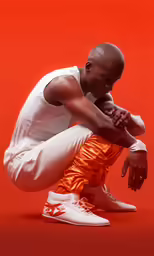 a young black man wearing a white shirt and orange pants crouching down with his legs folded