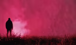 a man stands on a foggy hillside as pink light from the fog lifts up