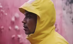 a man wearing a yellow hoodie against a pink wall