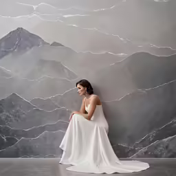 a woman in a white dress sits on a grey backdrop