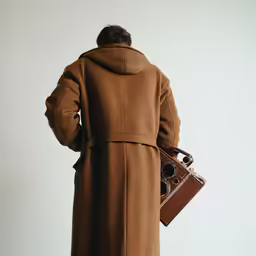 a man in a trench coat standing with his back turned, carrying an old camera