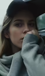 a woman wearing a baseball cap and a sweatshirt holding her hands on the front of her head while looking at something with