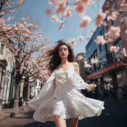woman in a white dress walking down the street with blossoming trees behind her
