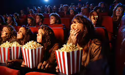 some women are in the movie seat looking up at the screen,some people are watching a movie and they are eating popcorn