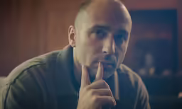 a bald man with a beard looks pensive while holding his finger to the temple